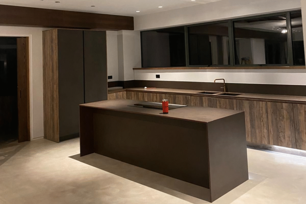 Modern kitchen interior featuring a sleek, dark wood island and cabinetry, with a minimalist design and large windows for natural light.