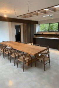 Modern dining area featuring a long wooden table with multiple chairs, contemporary kitchen design, large windows providing natural light, and a clean, minimalist aesthetic, reflecting Solved Construction's expertise in renovations and interior design.